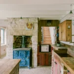 Kitchen at Purbeck Cottage by TYPE, restored blue cast-iron stove built into Purbeck stone chimney breast, reclaimed pine cabinetry, salvaged stone sink with brass taps