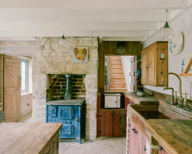 Kitchen at Purbeck Cottage by TYPE, restored blue cast-iron stove built into Purbeck stone chimney breast, reclaimed pine cabinetry, salvaged stone sink with brass taps