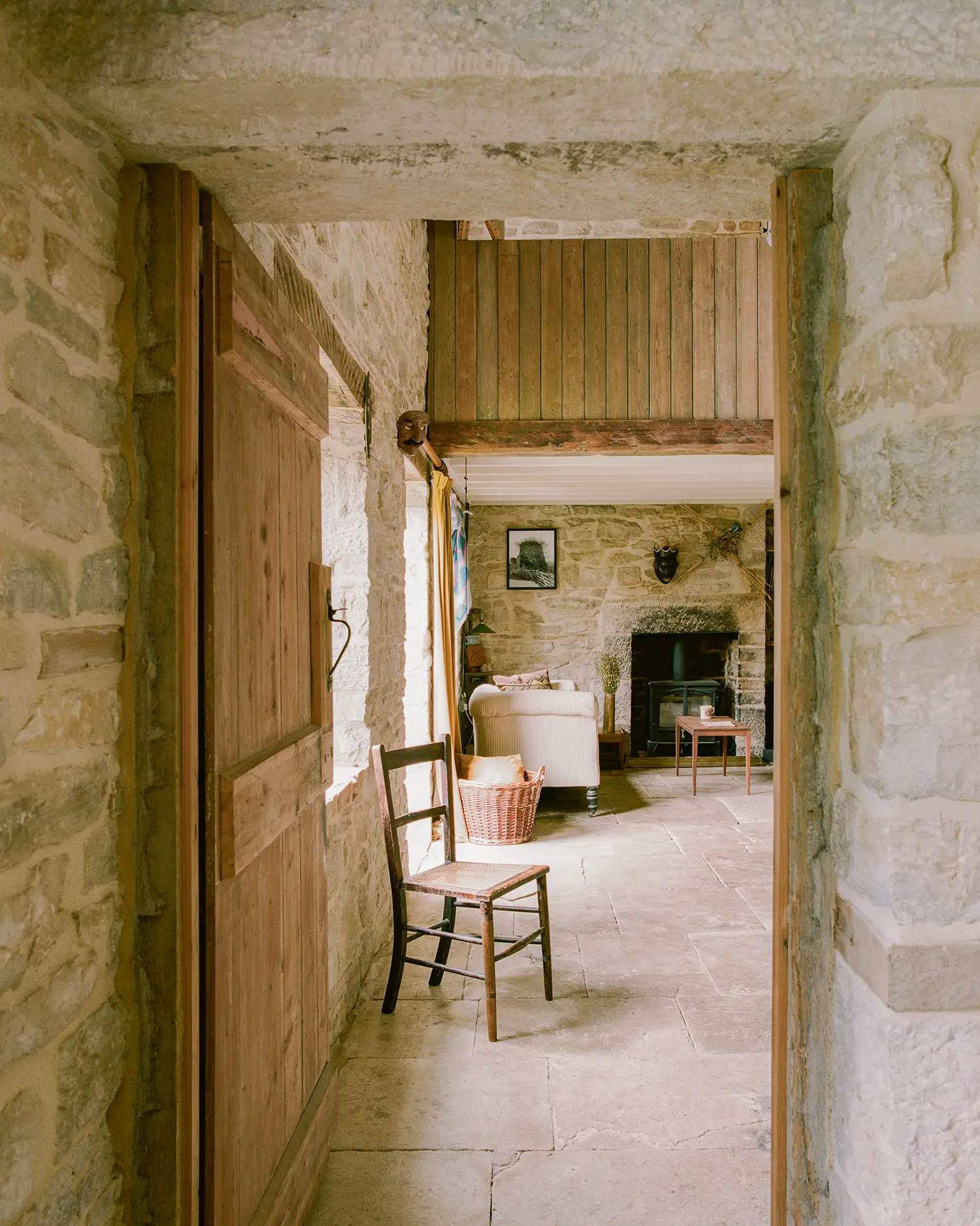 Interior threshold at Purbeck Cottage by TYPE, reclaimed timber door opening onto stone-floored living room with wood-burning stove and exposed stone walls