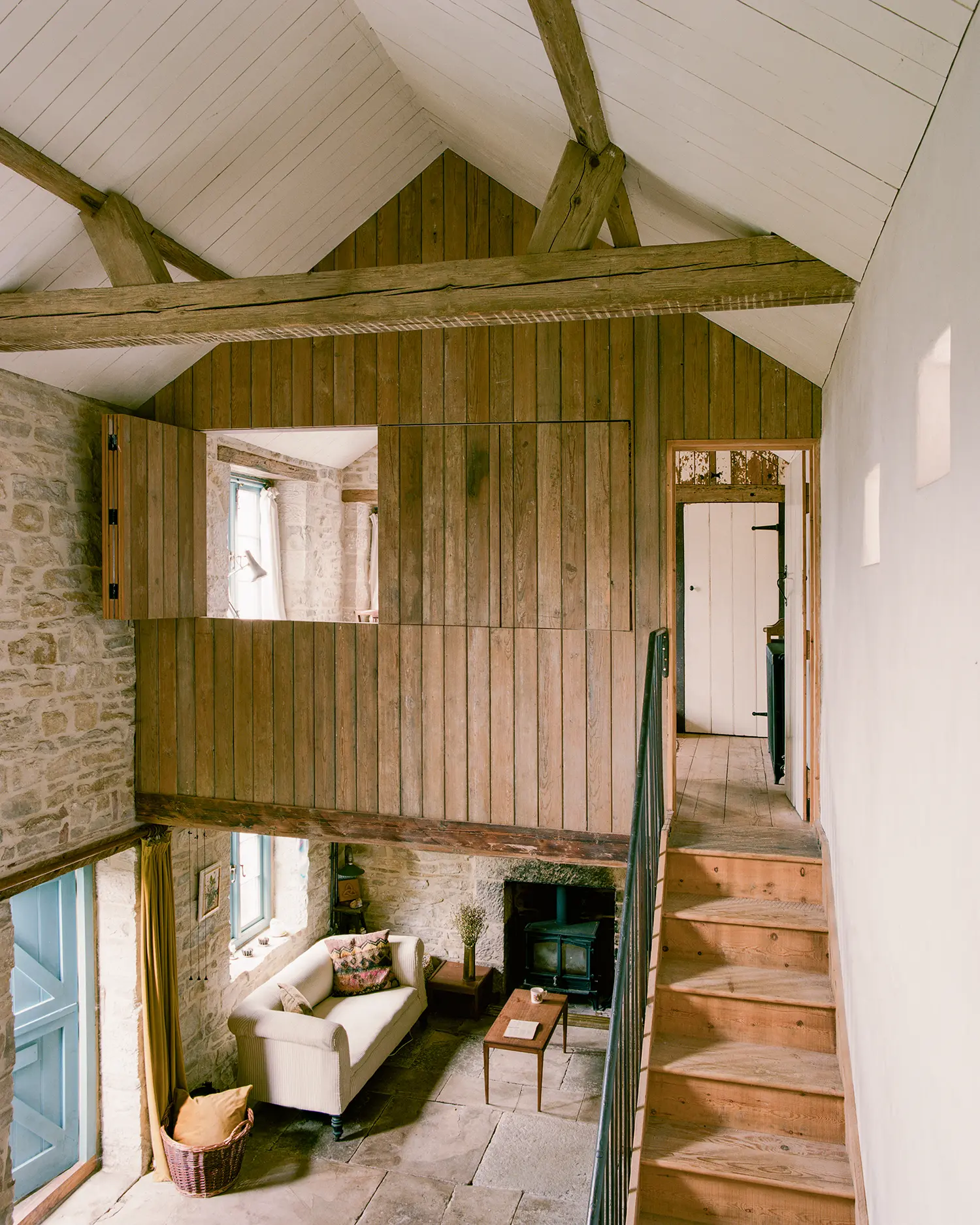 Double-height living space at Purbeck Cottage by TYPE viewed from mezzanine gallery, exposed stone walls, timber-clad volume, wood-burning stove below