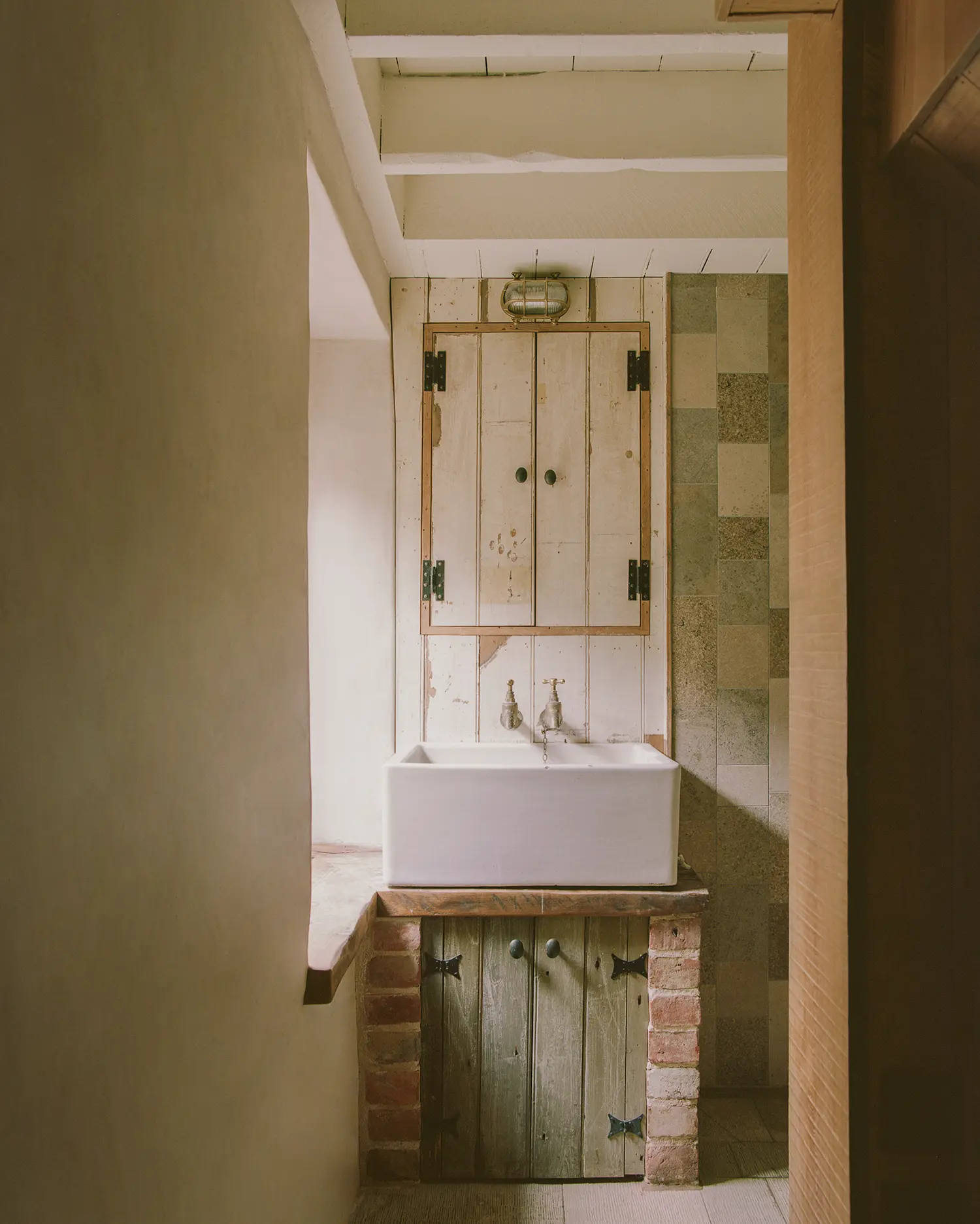 Bathroom at Purbeck Cottage by TYPE, salvaged ceramic Belfast sink on brick and timber stand beneath restored wall cabinet, patchwork stone tile behind