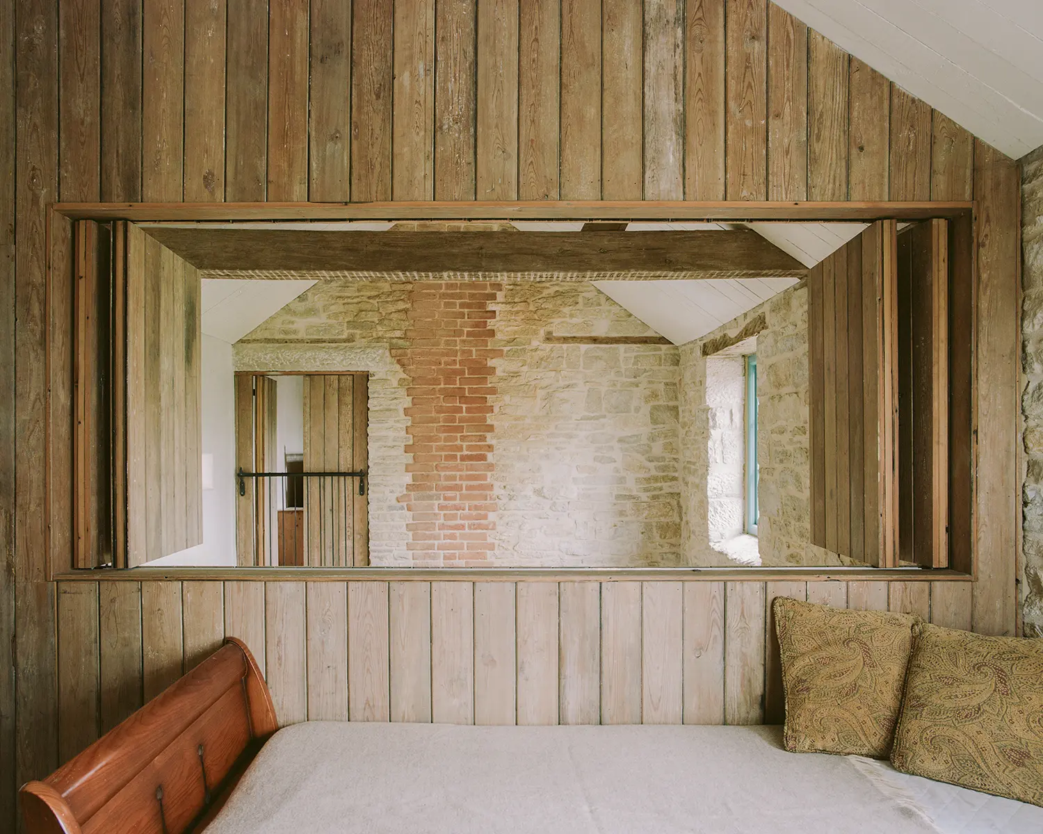 Bedroom at Purbeck Cottage by TYPE, salvaged timber wall paneling with bifold internal window opening onto exposed stone hallway and brick chimney breast