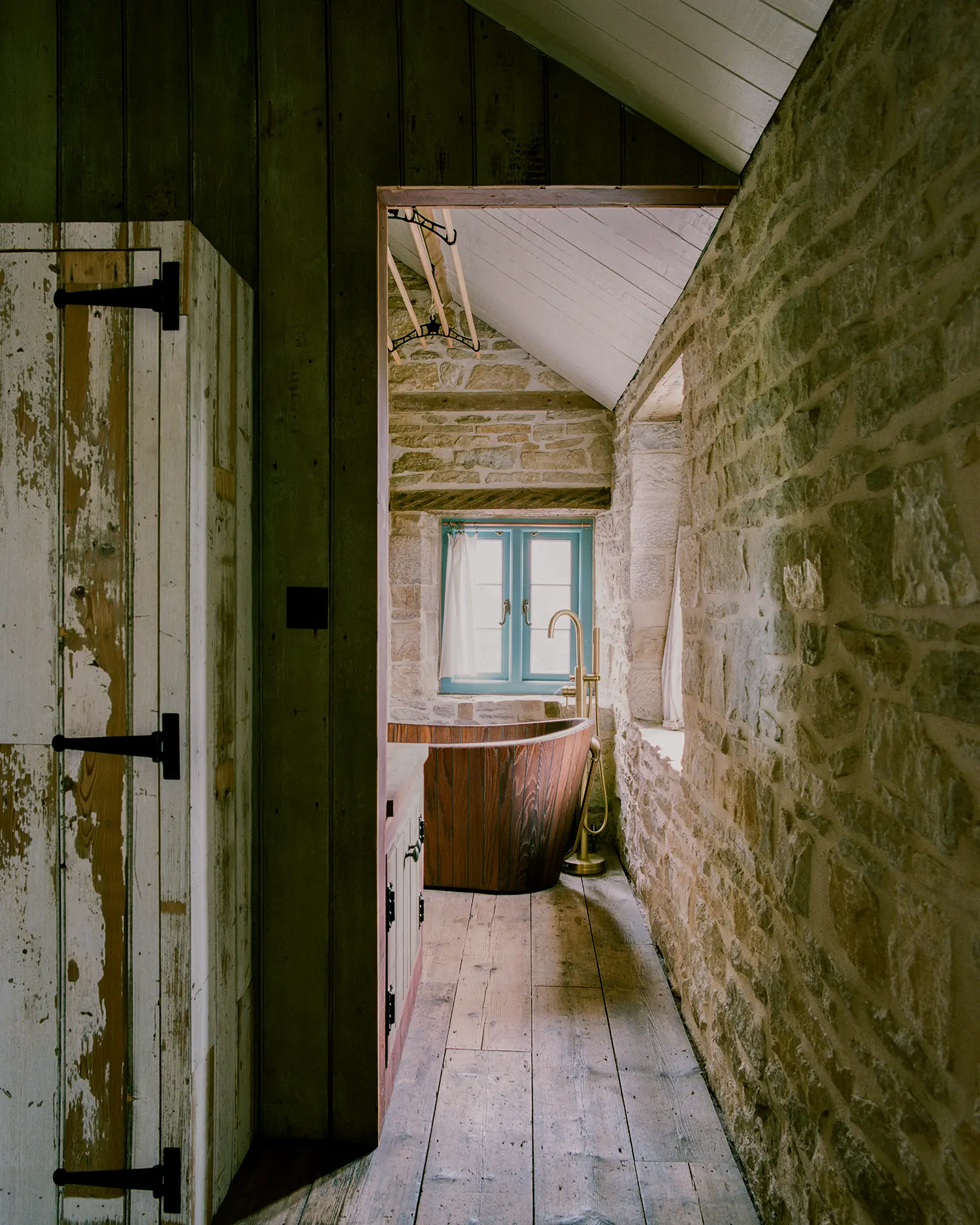 Attic bathroom at Purbeck Cottage by TYPE, freestanding wooden bathtub beneath a teal-painted stone window, exposed stone walls along a narrow corridor
