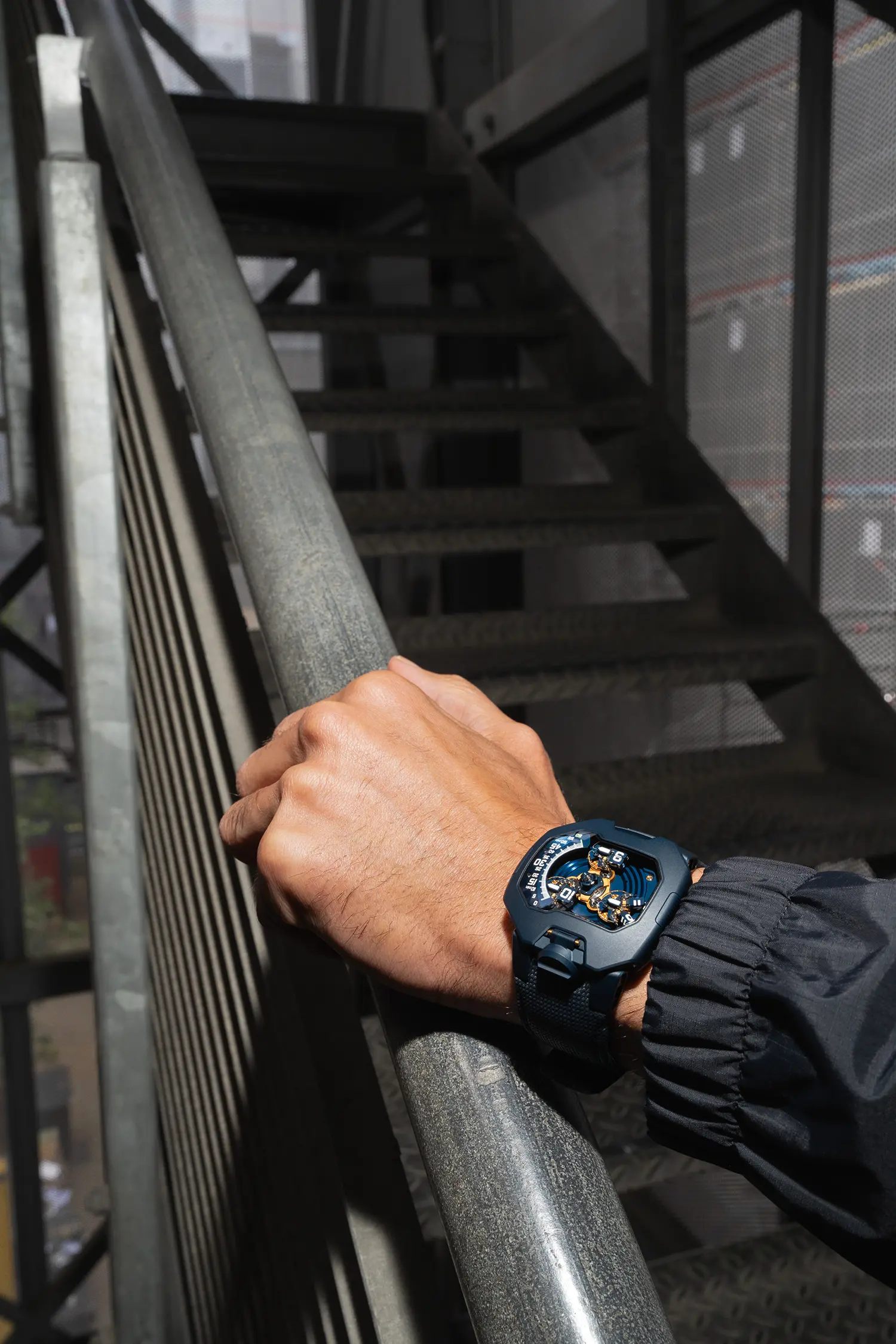 URWERK UR-120 Blue Planet on wrist gripping steel staircase railing, sandblasted blue case, blue Cordura strap, urban industrial outdoor setting