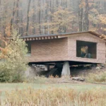 The Findling by Of Possible in Austerlitz, New York, larch-clad cabin raised on a concrete pier above a stone stair in autumn forest
