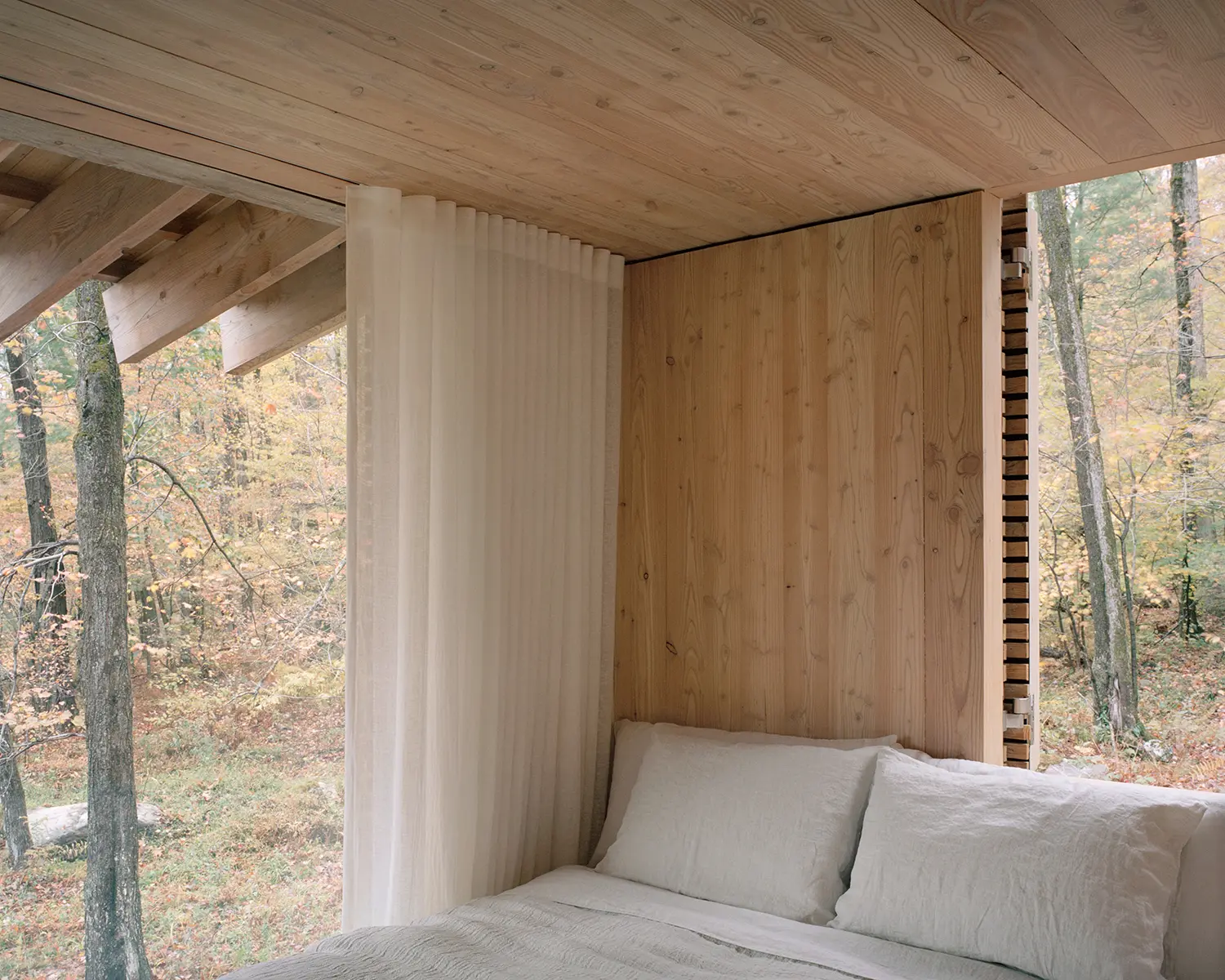 Corner bedroom nook in The Findling by Of Possible, linen bed against larch paneling with sheer curtain and forest view