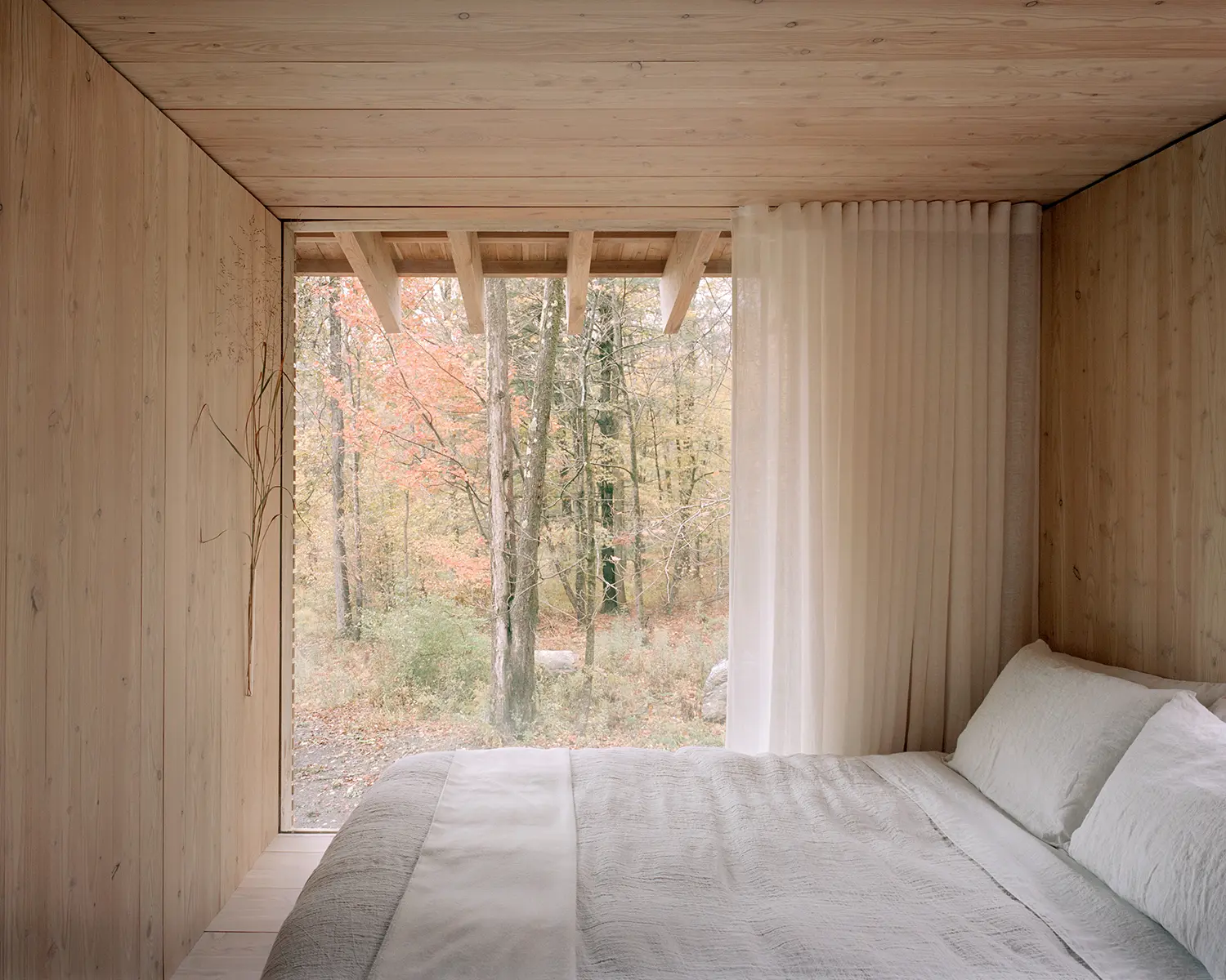 Bedroom window in The Findling by Of Possible, timber ceiling beams framing an autumn forest view beside a linen-covered bed