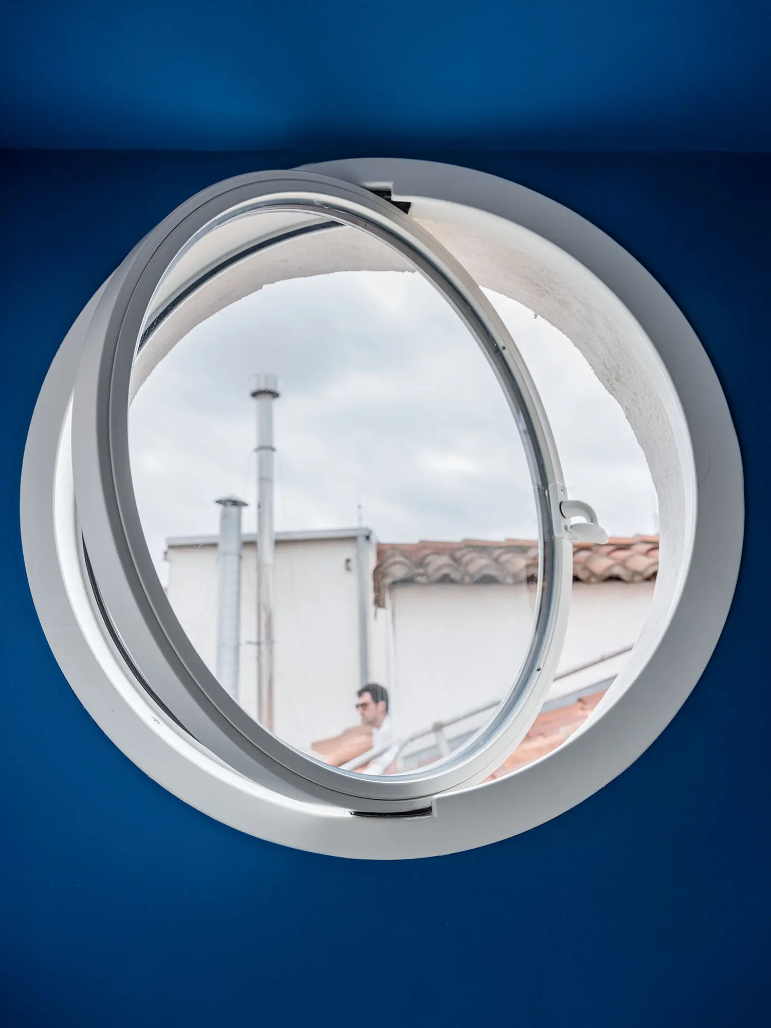 Close-up of open round porthole window against blue wall, view of rooftop chimneys, Madrid attic