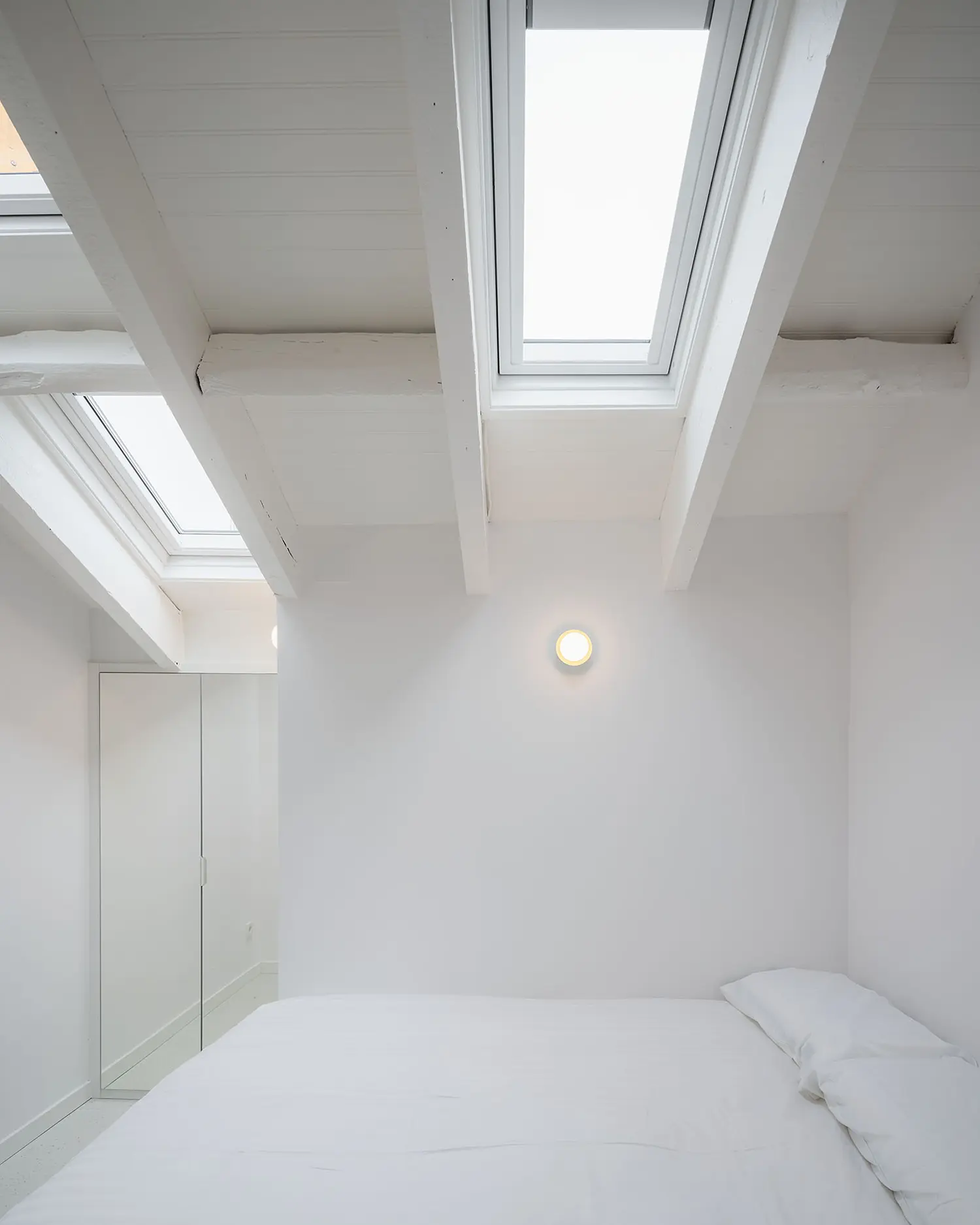 All-white attic bedroom with two vertical roof windows, exposed painted beams, minimal interior