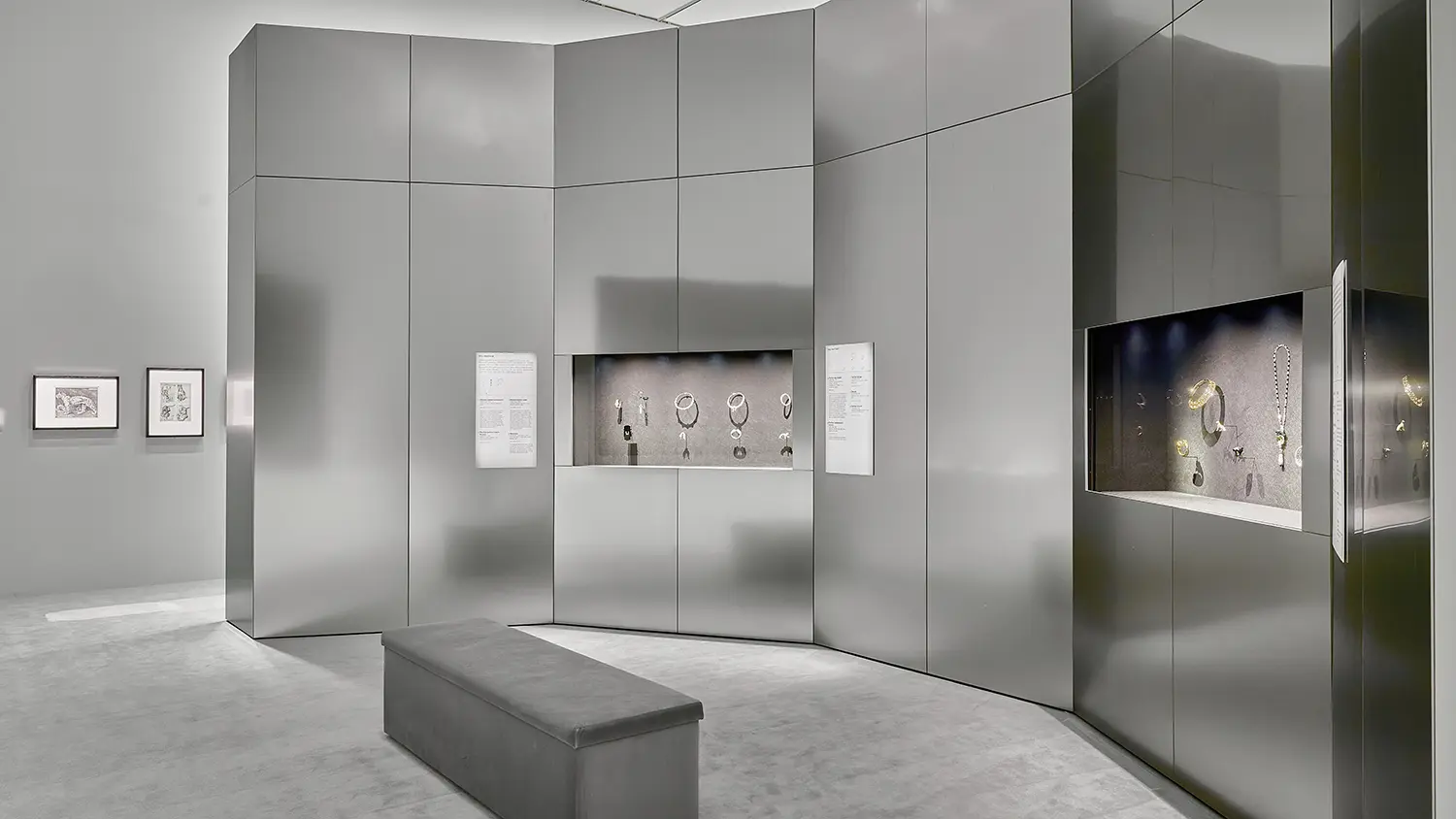 CARTIER exhibition NGV Melbourne: silver-grey curved gallery with gloss panel walls and recessed jewelry vitrines, Cartier archival drawings on the wall