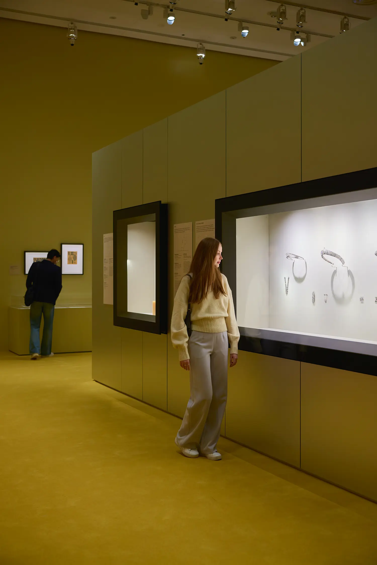 CARTIER at NGV Melbourne: olive-yellow gallery with visitor studying Cartier design drawings in wall-embedded illuminated vitrines