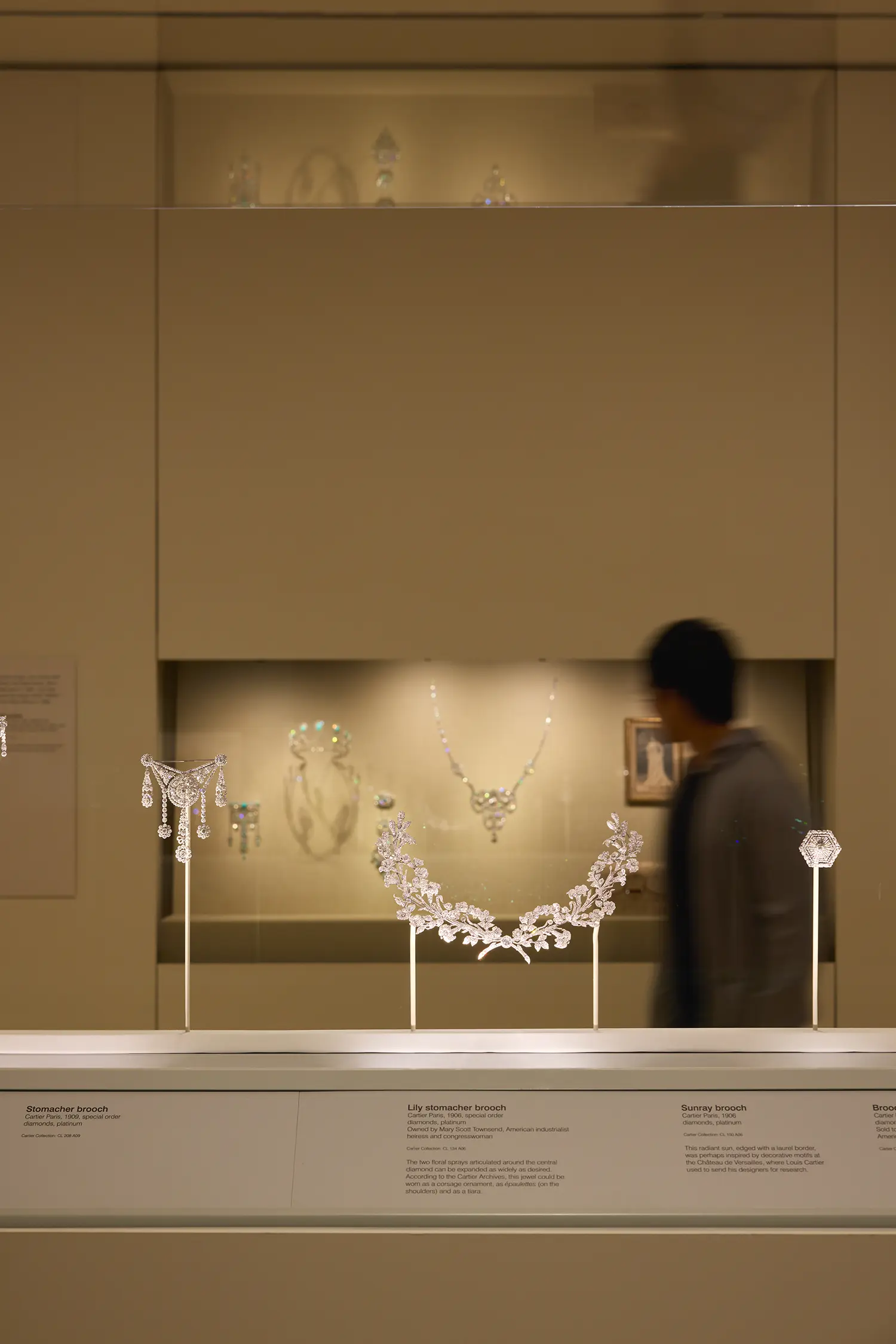 CARTIER exhibition NGV Melbourne: Garland Style diamond brooches and stomacher on display mounts in illuminated vitrine, visitor in motion behind