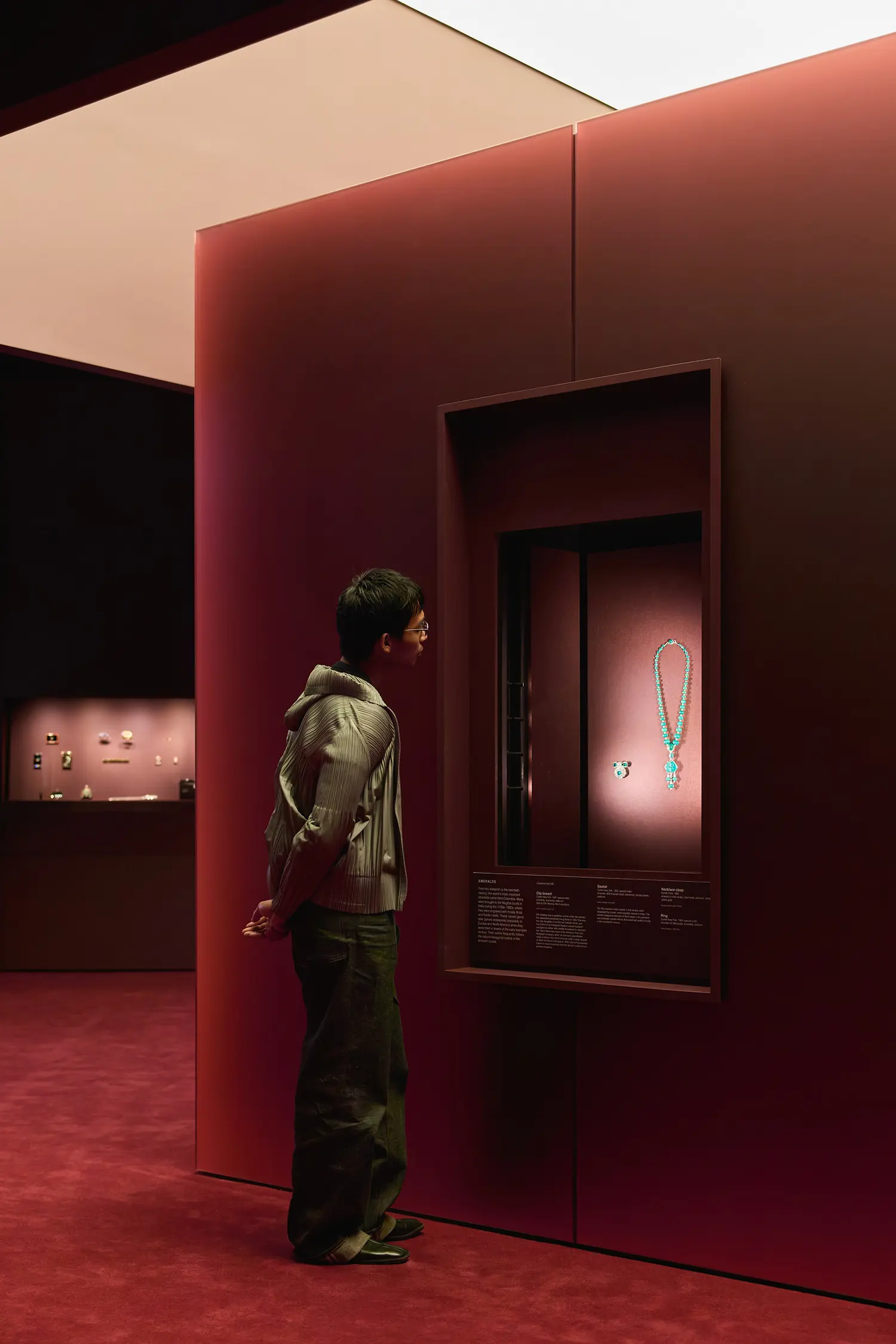 CARTIER at NGV Melbourne: visitor studying a turquoise necklace in a deep burgundy wall niche vitrine, jewelry cases visible in background