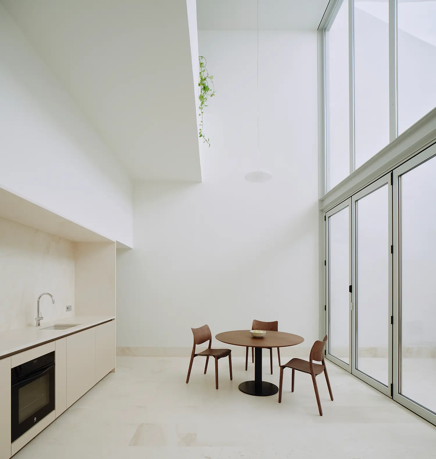 Ground-floor kitchen and dining area of Casa Nido by Iterare Arquitectos, double-height courtyard window, round wood table and three chairs