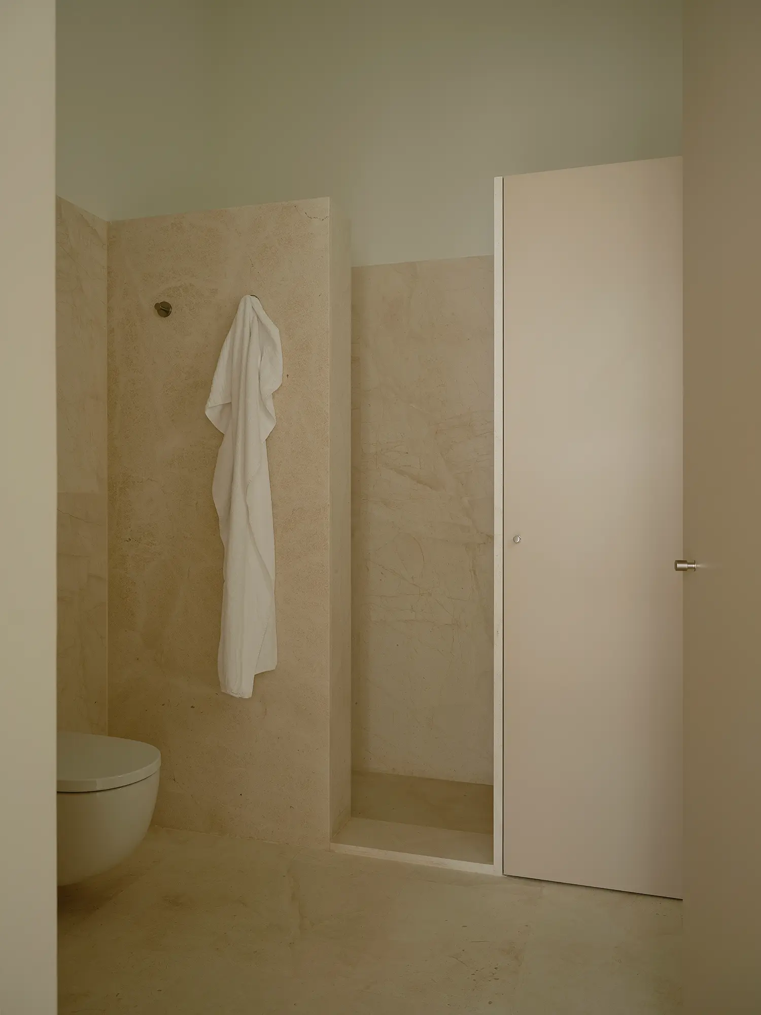 Bathroom in Casa Nido by Iterare Arquitectos, limestone-clad shower niche, wall-hung toilet and a towel hung on a hook