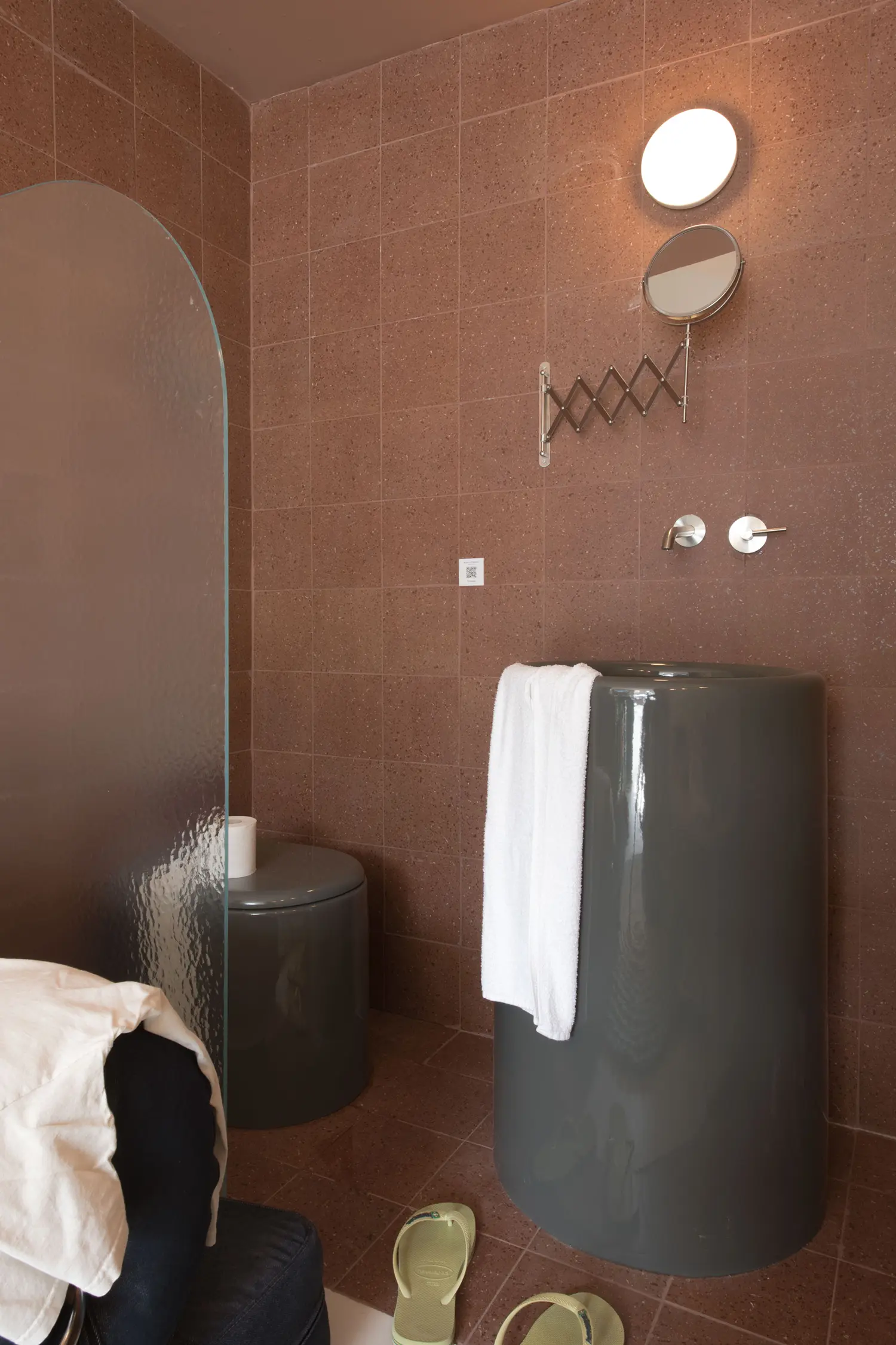 Container 03 by Atelierzero: terracotta-tiled bathroom with cylindrical ceramic sink, arched glass shower divider, and folded towel