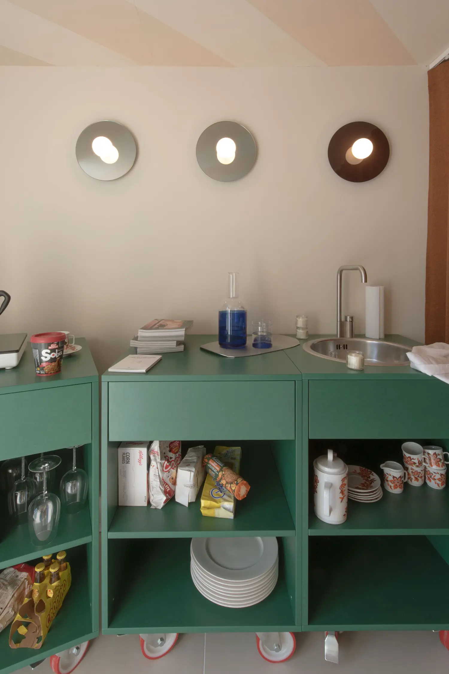 Container 03 installation, Atelierzero: mobile sage-green kitchen modules with sink, glassware, and packaged groceries on open shelving