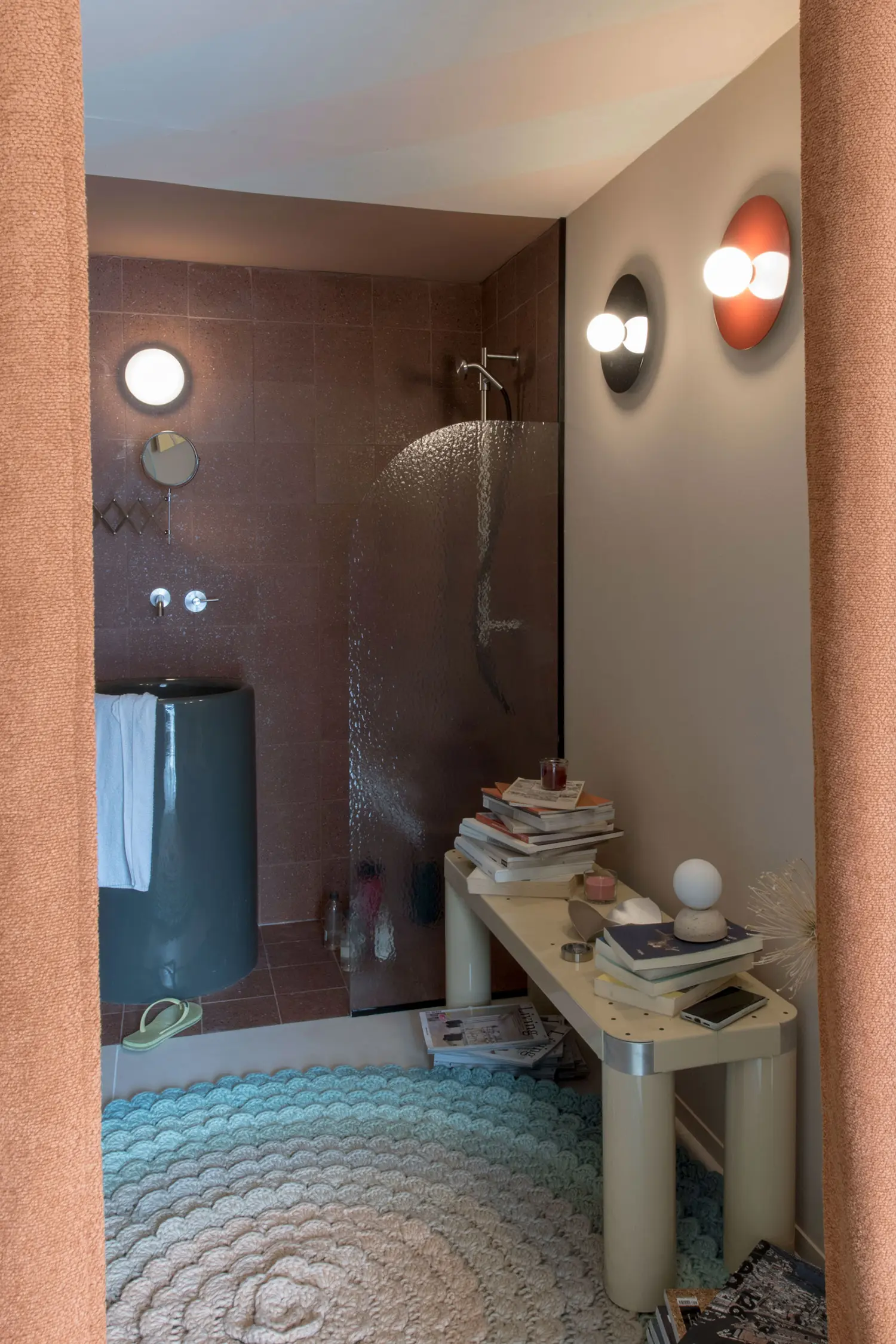 Container 03 by Atelierzero: terracotta bathroom with rain shower, cylindrical sink, cream tubular bench stacked with books, and blue rug