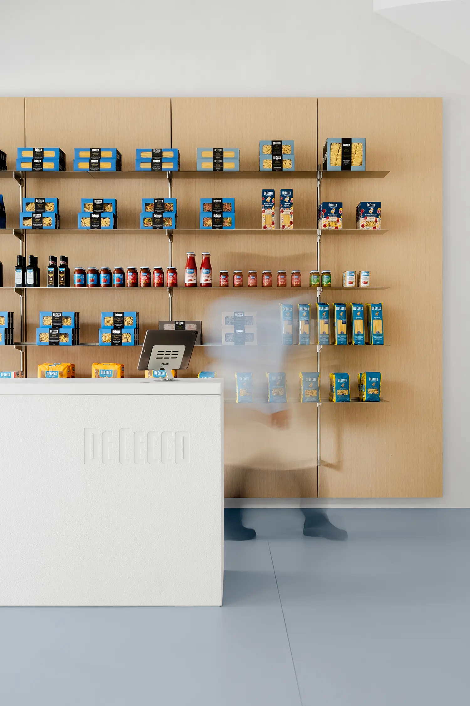 De Cecco Flagship Store by Studio Francesco Faccin, shelving wall with packaged pasta and sauces, blurred figure in motion near checkout counter