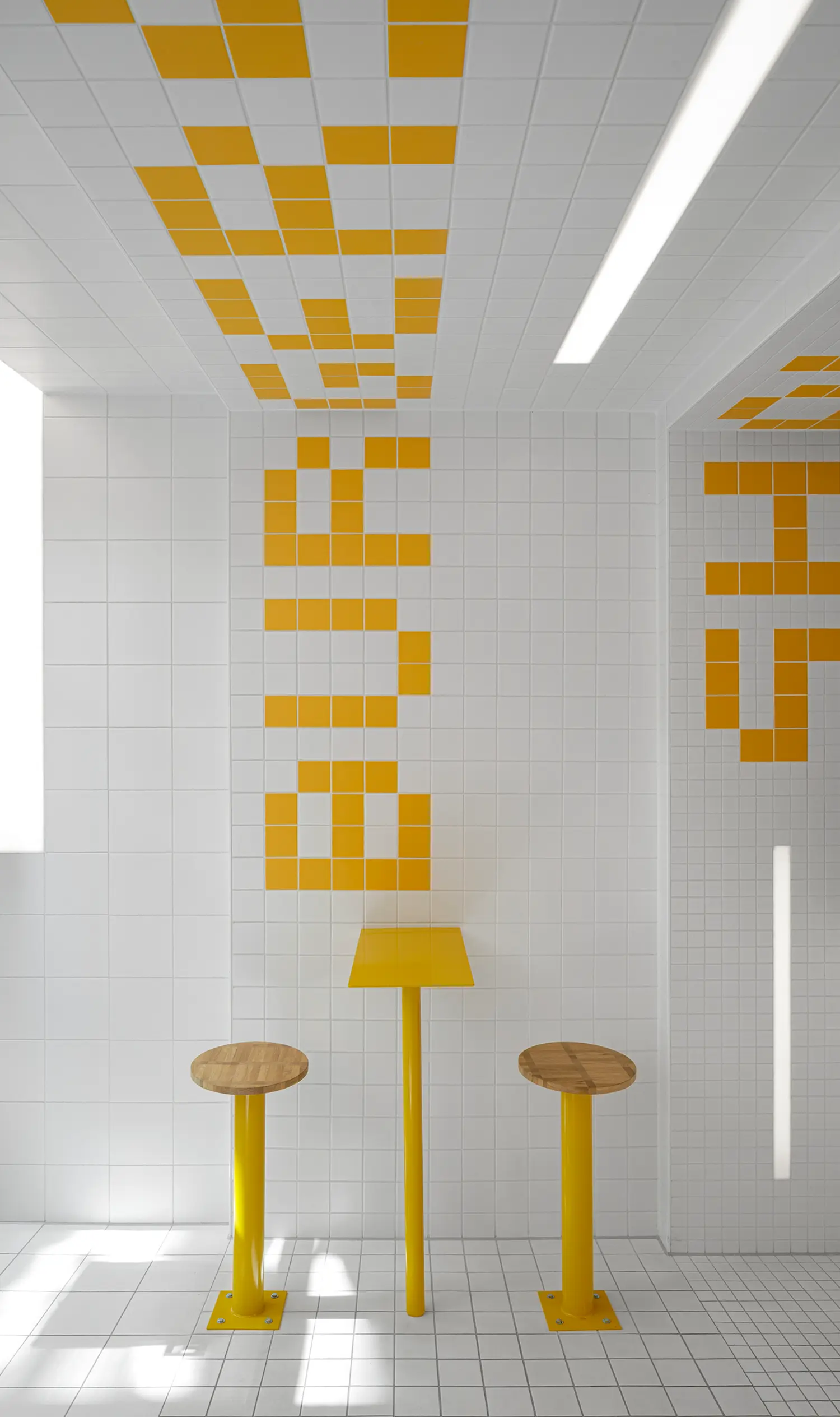 Angled view of yellow tile typography on ceiling and wall at Fab Fries Freddy by Ninetynine, with round table and wooden stools below