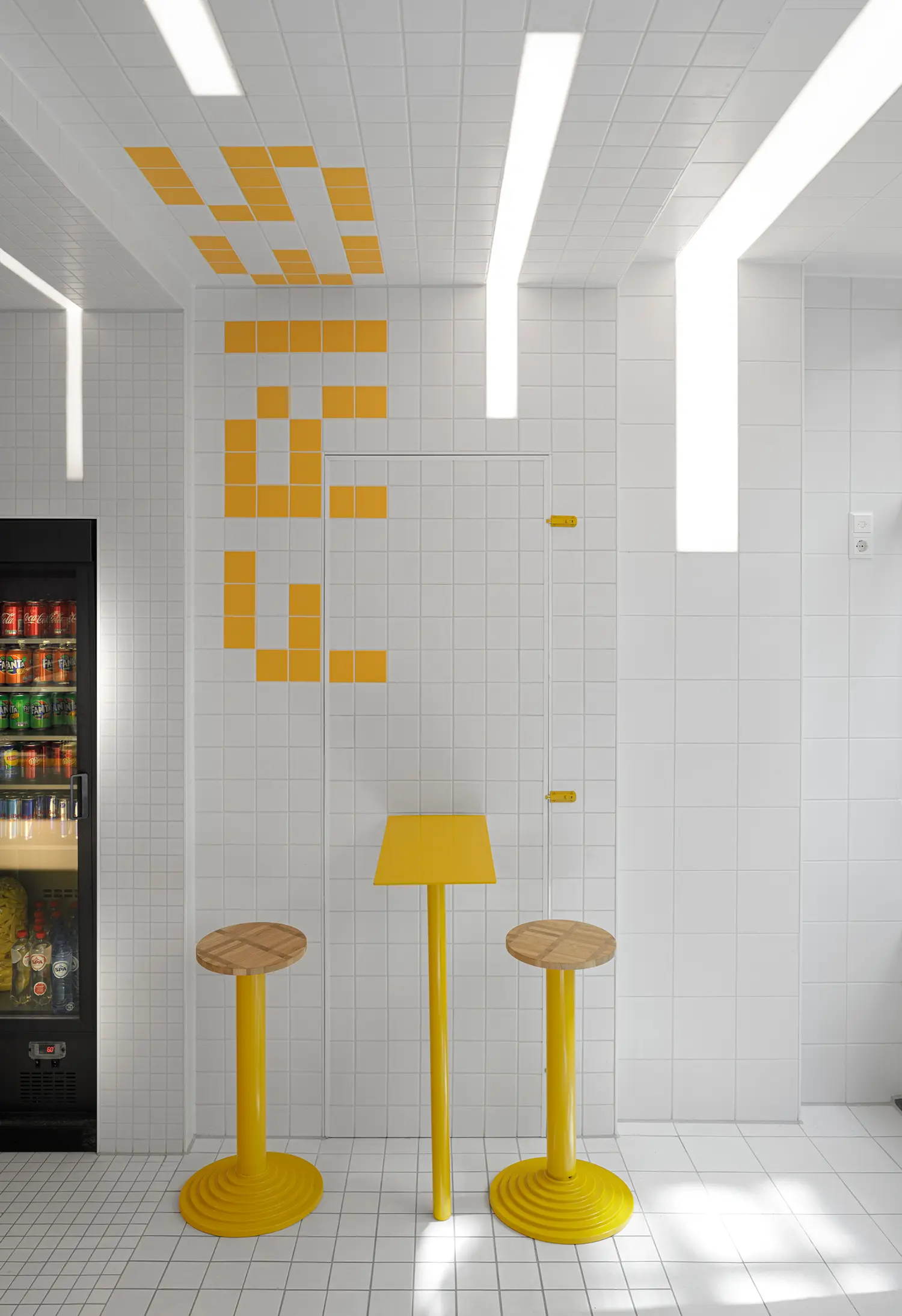 Interior detail of Fab Fries Freddy by Ninetynine, Amsterdam, showing drinks fridge, glass door with yellow hardware, and round yellow table