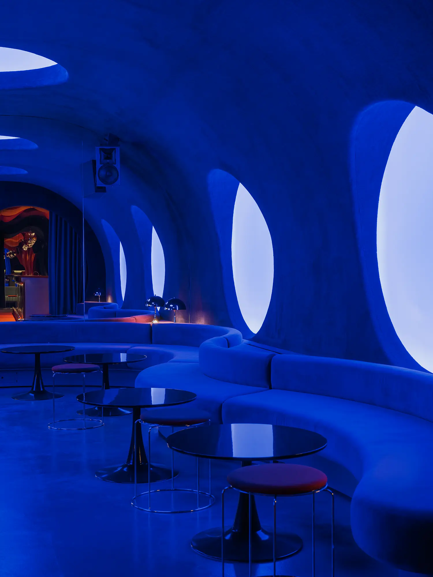 Blue DJ room in Focacha by Isern Serra, oval illuminated wall openings above a curved banquette and round tables with red stools