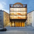 Kinderoase an der TUM by Kéré Architecture, five-story timber kindergarten with slatted wood facade lit at dusk between two older Munich buildings