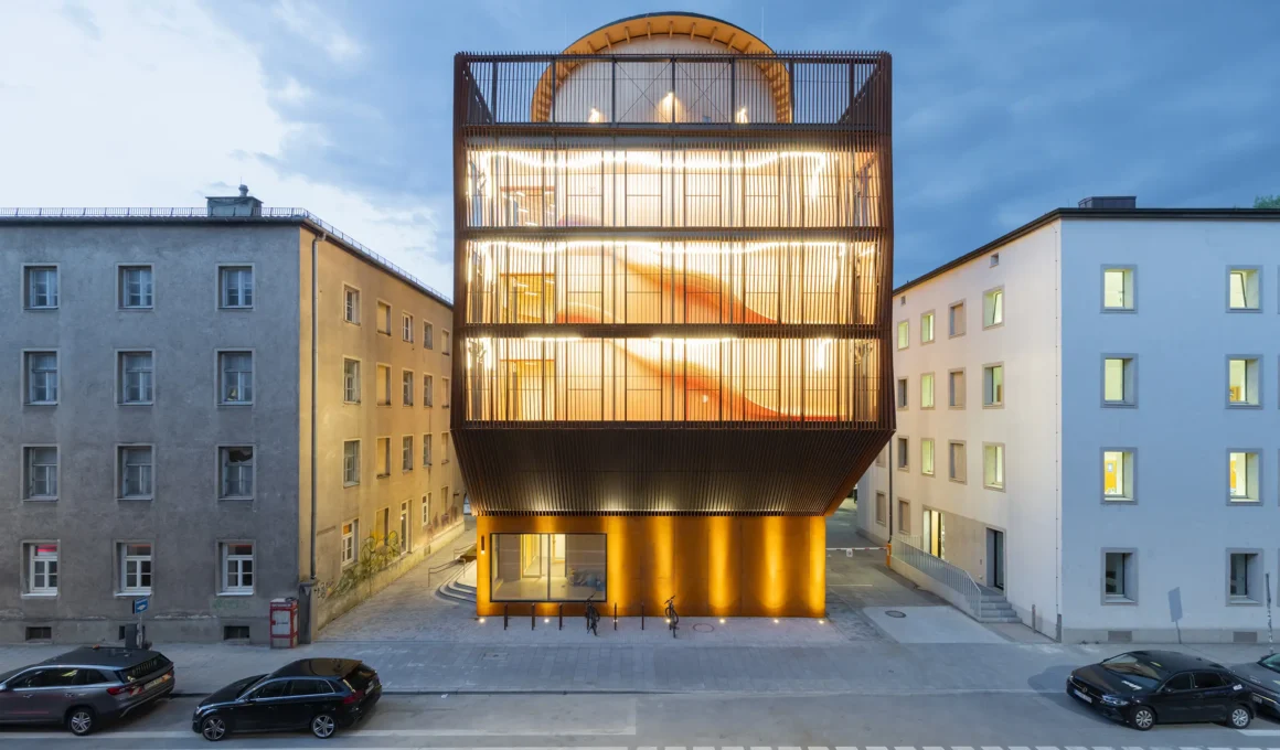 Kinderoase an der TUM by Kéré Architecture, five-story timber kindergarten with slatted wood facade lit at dusk between two older Munich buildings