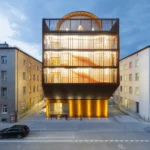 Kinderoase an der TUM by Kéré Architecture, five-story timber kindergarten with slatted wood facade lit at dusk between two older Munich buildings