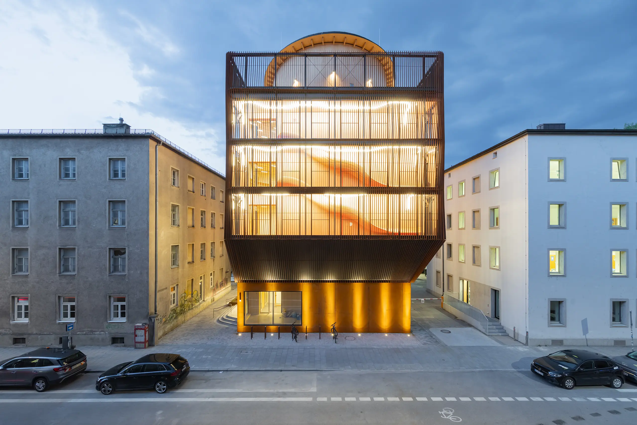 Kinderoase an der TUM by Kéré Architecture, five-story timber kindergarten with slatted wood facade lit at dusk between two older Munich buildings