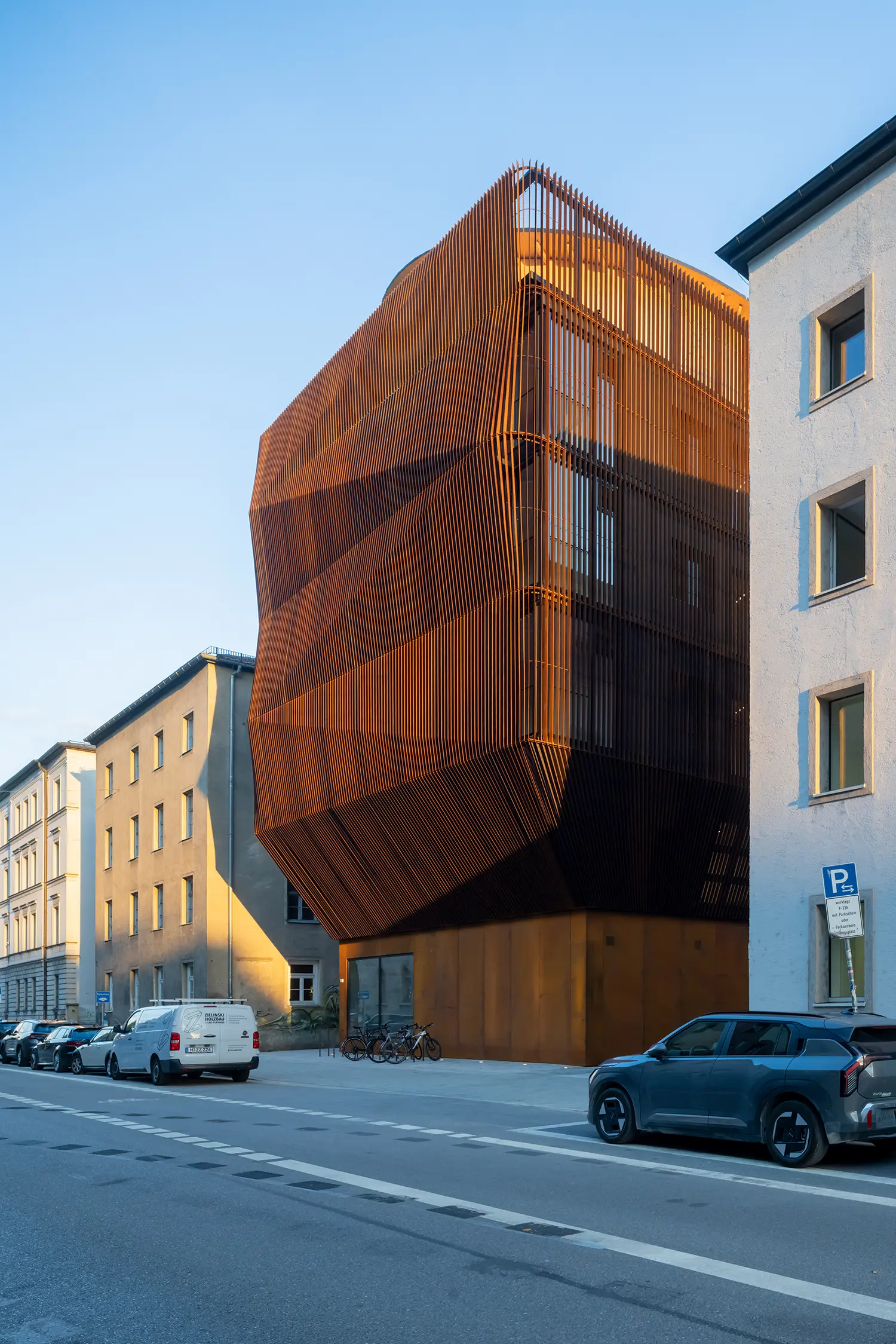 Corner view of the Kinderoase an der TUM by Kéré Architecture, faceted timber-slat volume catching low sun above a Munich street
