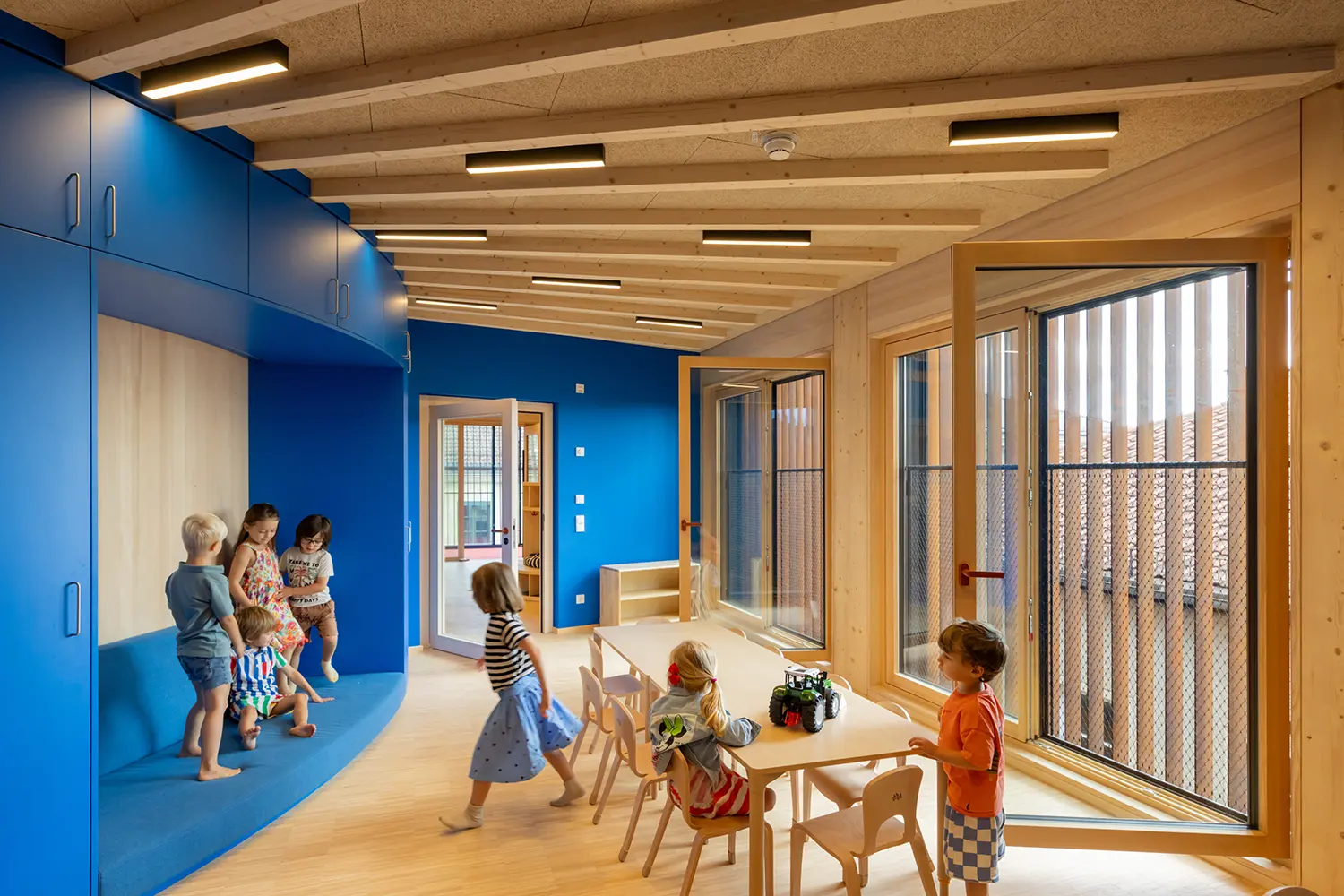 Blue-walled classroom in Kéré Architecture's timber kindergarten, children at a low table beside timber-beam ceiling and slatted windows