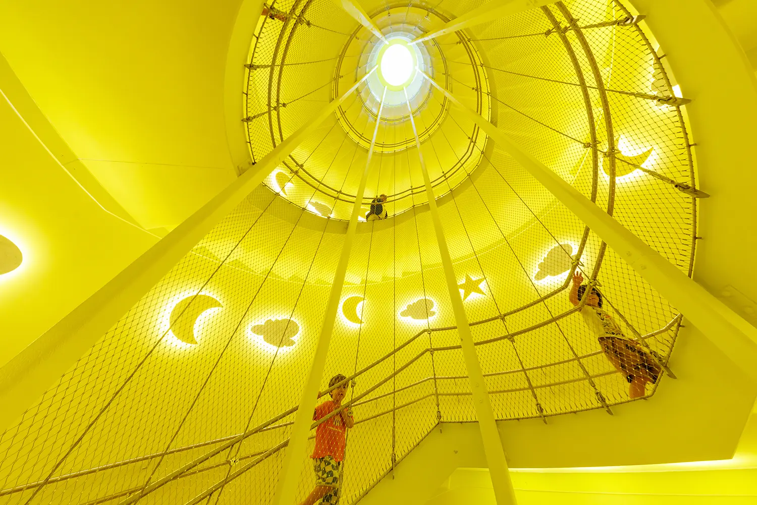 Yellow-lit rooftop play dome inside the Kinderoase an der TUM by Kéré Architecture, children climbing safety netting under moon and star cutouts