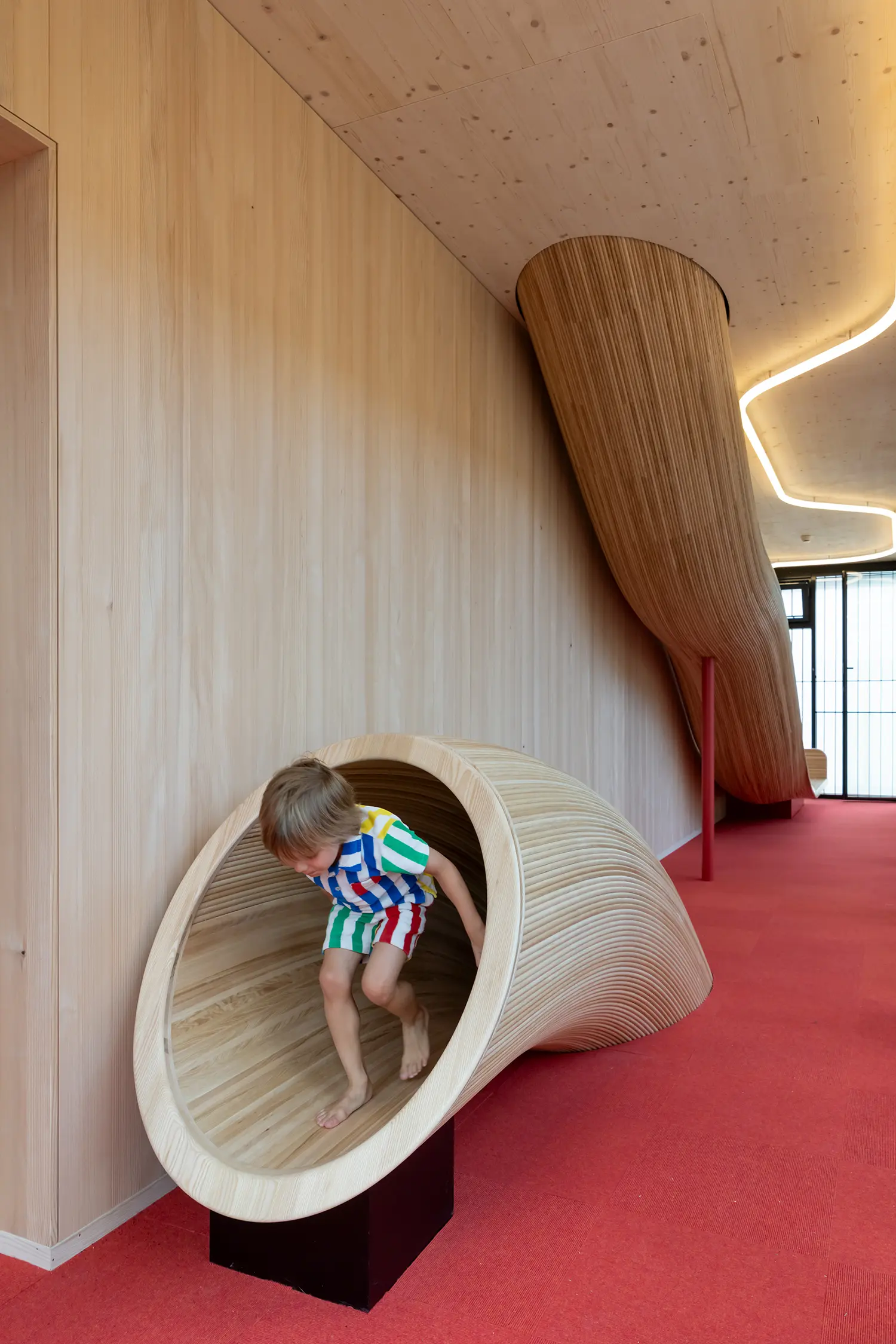 Child climbing into a sculptural timber slide pod in Kéré Architecture's kindergarten, curved wood form against a red floor