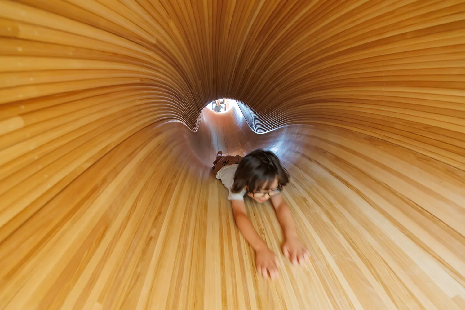 Interior view down a curved timber slide in Kéré Architecture's kindergarten, a child sliding through the wooden tube in motion