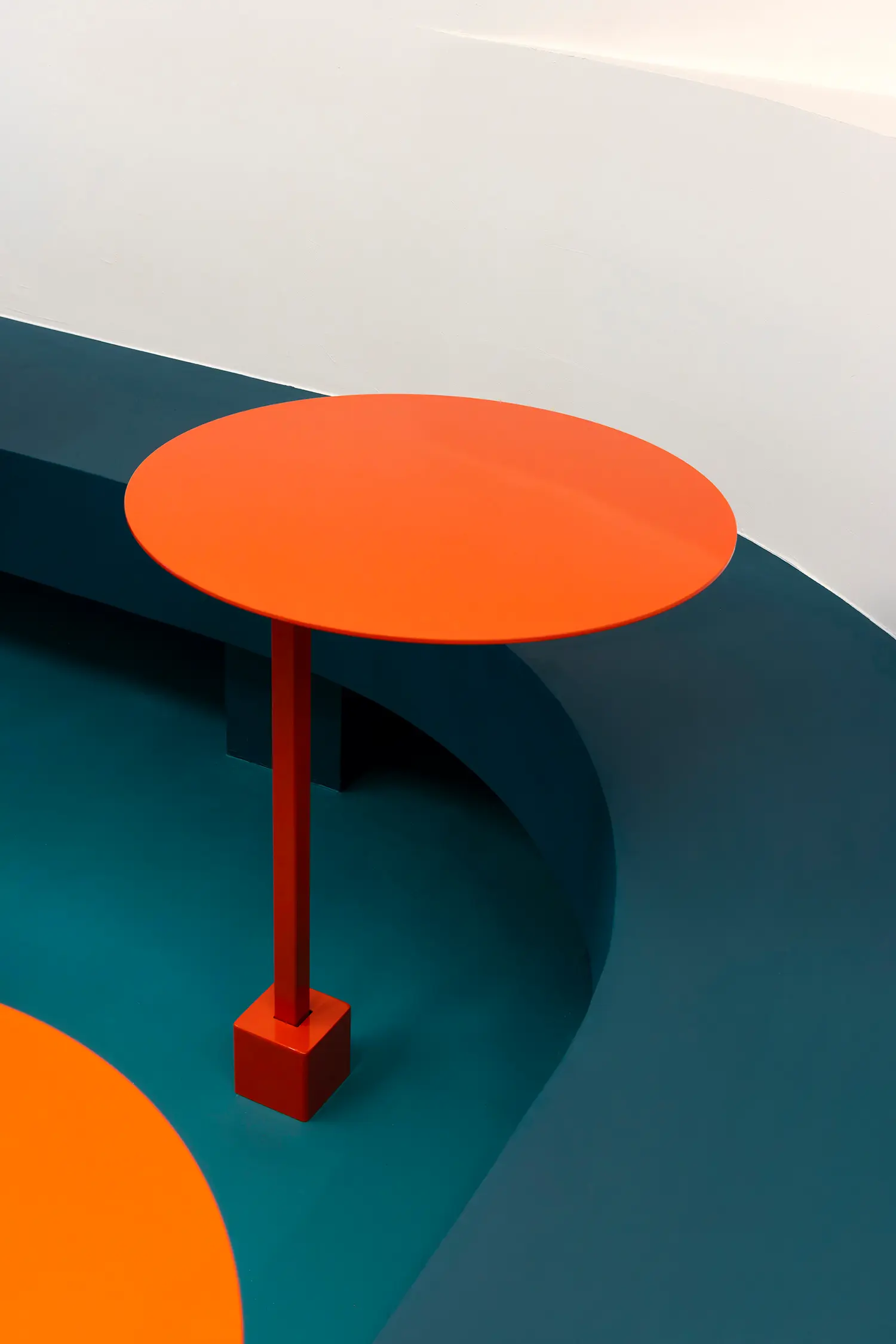 Sainto by Mur Mur, detail of red round table top on cube base against teal curved floor