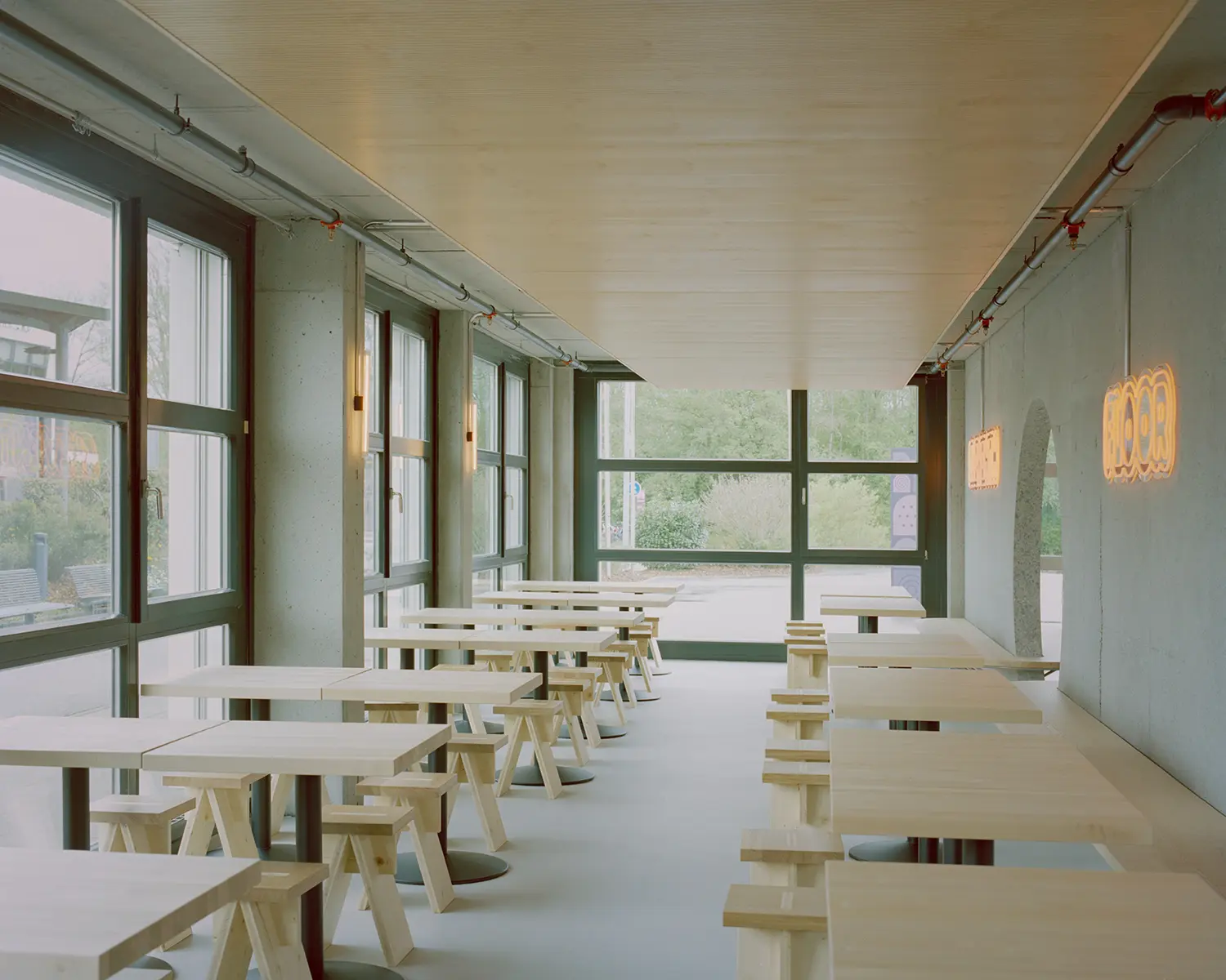 Eleventhfloor Restaurant by Noue Studio, long dining hall with wood tables, stools, and concrete archway with neon signage