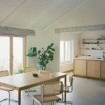 Dining area with oak table, cane chairs, and open kitchen beyond concrete lintel in Noue Studio villa