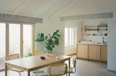Dining area with oak table, cane chairs, and open kitchen beyond concrete lintel in Noue Studio villa