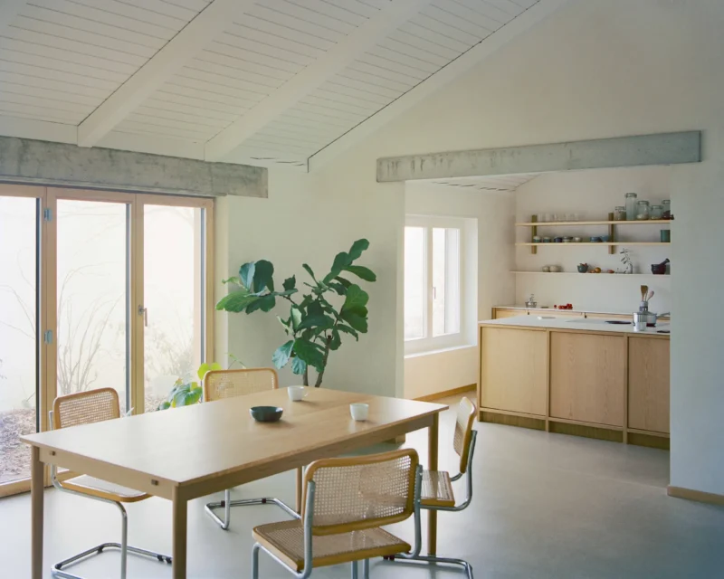 Dining area with oak table, cane chairs, and open kitchen beyond concrete lintel in Noue Studio villa