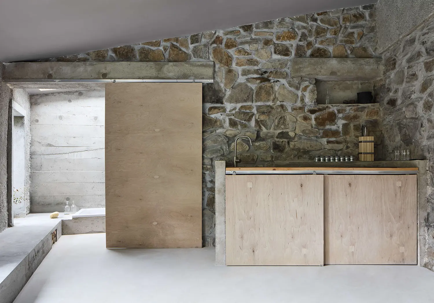 Panoší Újezd Summer House by Lenka Milerová: cleaned original stone wall behind a plywood kitchenette with concrete shelf and sliding plywood door