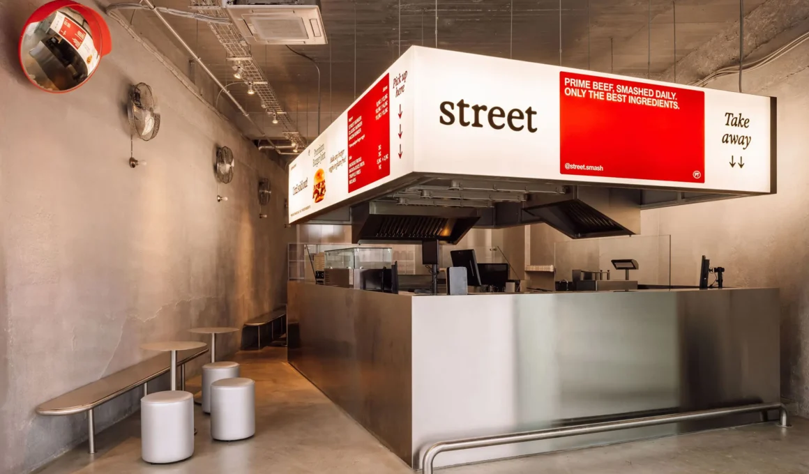 Wide interior view of Street Smash Burgers by XXXI.studio showing the illuminated silver kiosk, open kitchen, and stool seating in Almada