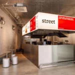 Wide interior view of Street Smash Burgers by XXXI.studio showing the illuminated silver kiosk, open kitchen, and stool seating in Almada