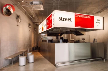 Wide interior view of Street Smash Burgers by XXXI.studio showing the illuminated silver kiosk, open kitchen, and stool seating in Almada
