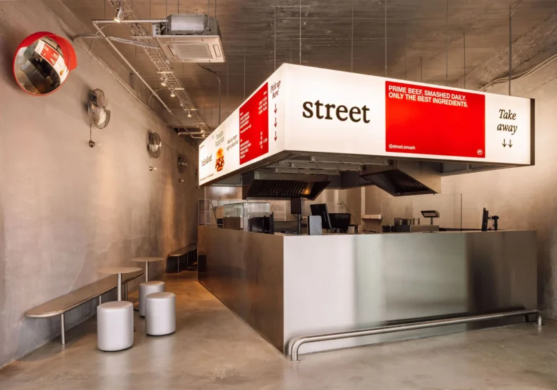 Wide interior view of Street Smash Burgers by XXXI.studio showing the illuminated silver kiosk, open kitchen, and stool seating in Almada