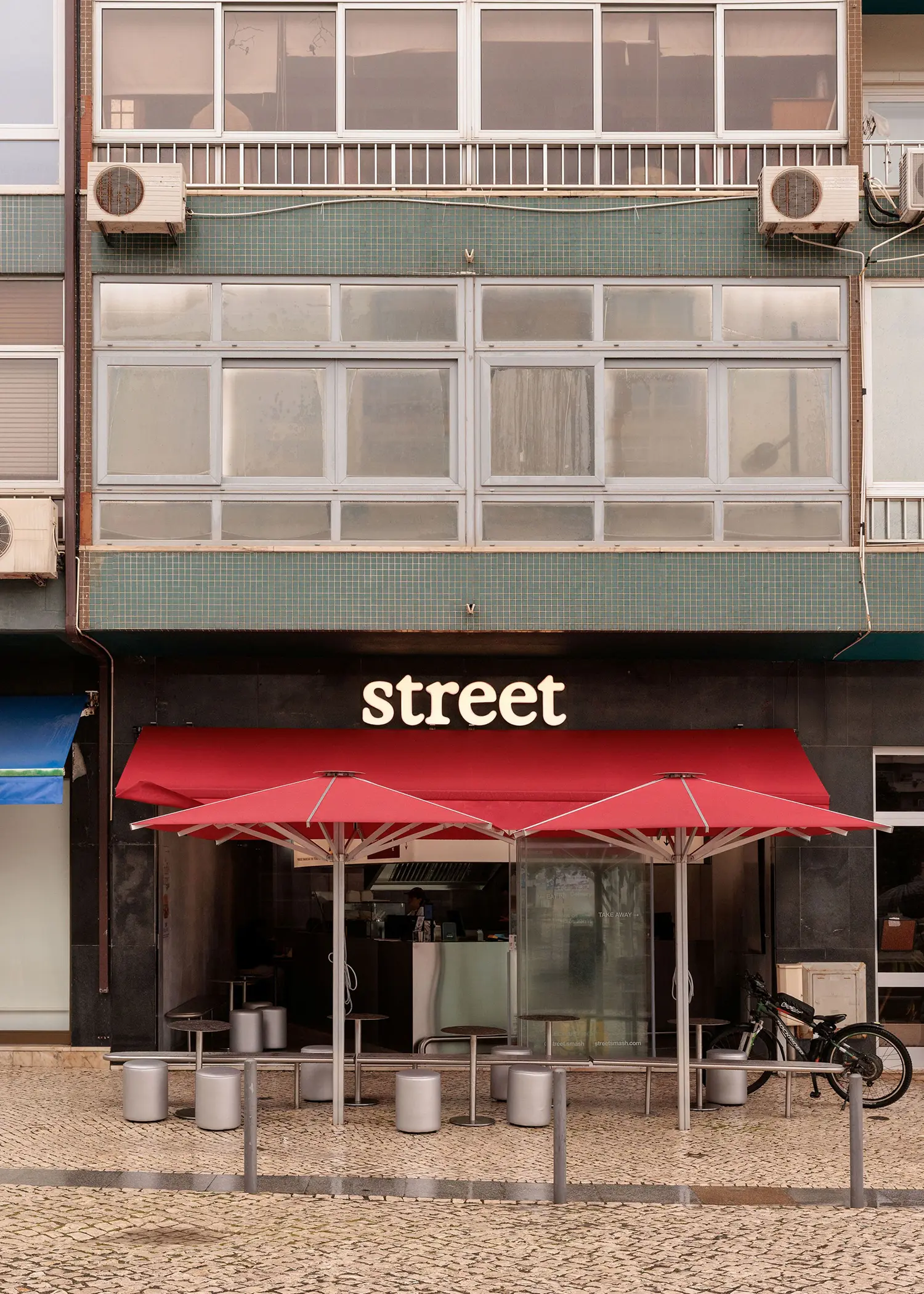 Exterior facade of Street Smash Burgers by XXXI.studio with red umbrella awnings on a public square in Almada, Lisbon