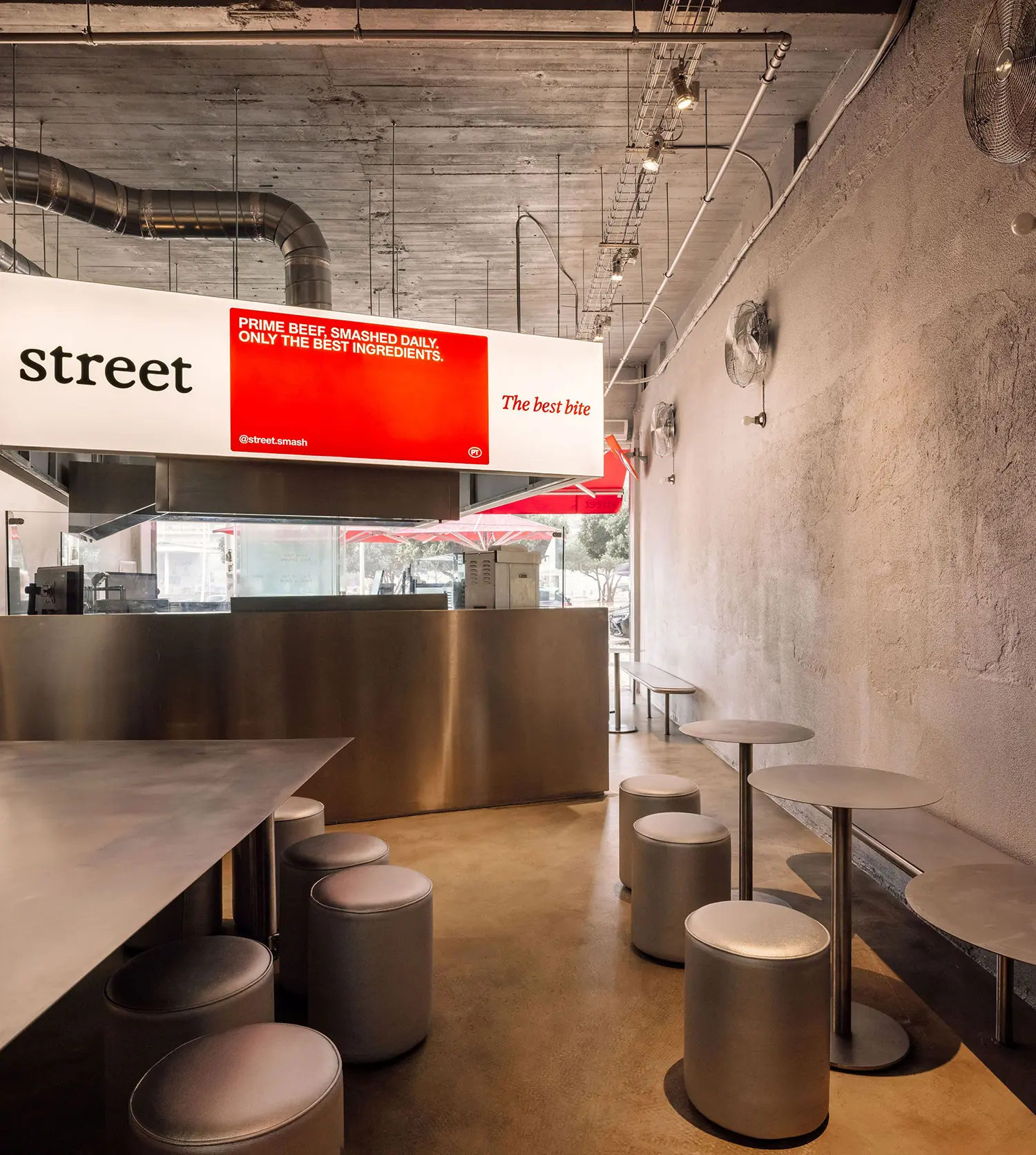 Interior seating area at Street Smash Burgers by XXXI.studio with red patio umbrellas visible through the storefront glass, Almada