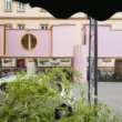 ZuHaus by Clemens Hoyer, pink timber structure on stilts above a Munich street, partially obscured by roadside greenery