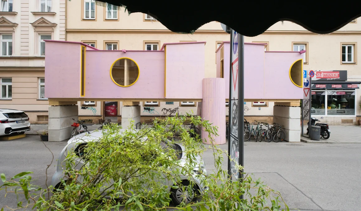 ZuHaus by Clemens Hoyer, pink timber structure on stilts above a Munich street, partially obscured by roadside greenery