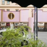 ZuHaus by Clemens Hoyer, pink timber structure on stilts above a Munich street, partially obscured by roadside greenery